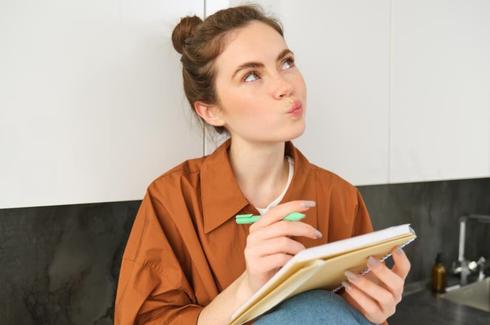 A young woman pensively jotting in a notebook in a kitchen